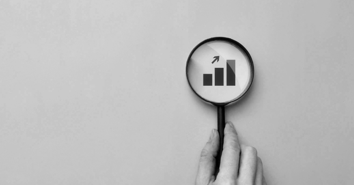 A hand holds a magnifying glass. Through the lens is a chart showing upward growth with an arrow pointing upwards.
